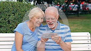 Senior couple having great time sitting on bench in park chatting relaxing, browsing in smartphone.