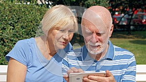 Senior couple having great time sitting on bench in park chatting relaxing, browsing in smartphone.