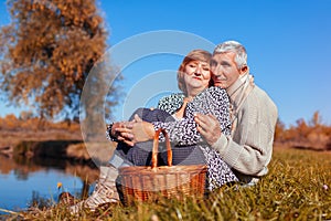 Senior couple having picnic by autumn lake. Happy man and woman enjoying nature and hugging