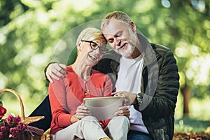 Senior couple having a picnic in park using digital tablet