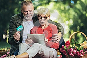 Senior couple having a picnic in park using digital tablet and credit card for online shopping