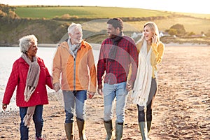 Senior Couple Hold Hands As They Walk Along Shoreline With Adult Offspring On Winter Beach Vacation