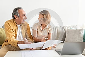 Senior couple holding documents reading paper bills, paying bank loan online, using laptop at home, free space