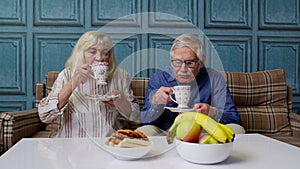 Senior couple holding drinking tea talking, laughing, smiling, kissing, sitting on couch at home