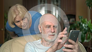 Senior couple at home. Husband using smartphone. Wife spying on her husband from his shoulder