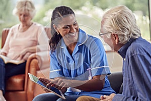 Senior Couple At Home With Man Talking To Female Nurse Or Care Worker Using Digital Tablet