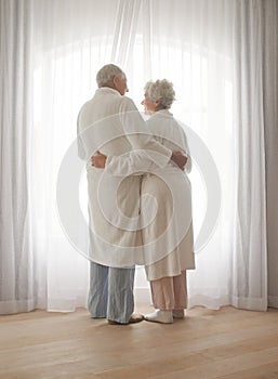 Senior couple, hug and relaxing by window in retirement, love and bonding in pajamas on holiday. Elderly people, back