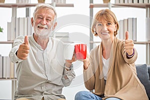 Senior couple inside home during a coffee break, Smiling elderly couple holding cups of coffee with showing thumbs up