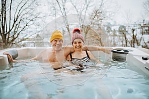 Senior couple in kintted cap enjoying together outdoor bathtub at their terrace during cold winter day. photo