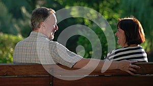 Senior couple laughing while sitting on bench in summer park.