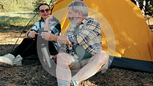 Senior couple, man and woman hiking in forest on warm day, sitting near tent and drinking hot tea