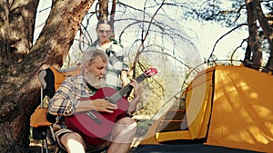 Senior couple, man and woman hiking in forest on warm day, sitting near tent. Man playing guitar