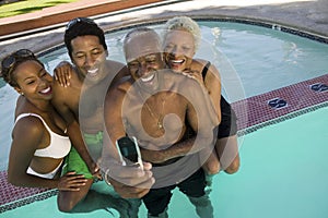 Senior couple and mid-adult couple posing for mobile phone photograph at swimming pool elevated view.