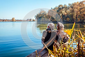 Senior couple relaxing by autumn lake. Happy man and woman enjoying nature and hugging sitting on pier