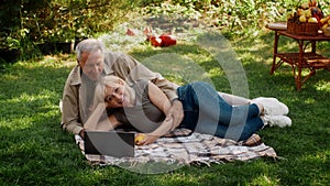 Senior couple relaxing with laptop outdoors, lying on picnic blanket in garden