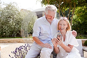 Senior Couple Relaxing Sitting Outdoors In Garden At Home Looking At Mobile Phone