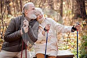 Senior couple resting while sitting on bench with nordic walking poles, smiling
