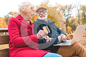 senior couple shopping online with laptop and credit card while sitting on bench