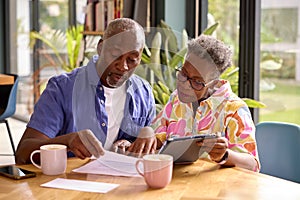 Senior Couple Sitting Around Table At Home Reviewing Finances Using Digital Tablet