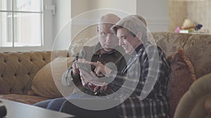 Senior couple sitting on the big sofa and bald man showing photos in the mobile phone for his woman with short grey hair