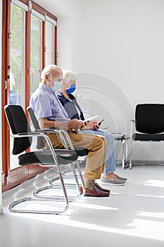 Senior couple sitting with face masks in a bright waiting room of  a hospital or an office