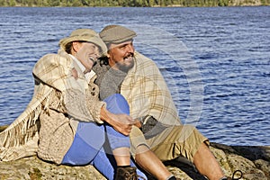 Senior couple sitting by a lakeside with blanket around shoulders.