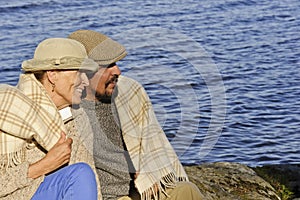 Senior couple sitting by a lakeside with blanket around shoulders.