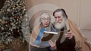 Senior couple spending happy time at home reading a book together with a decorated christmas tree