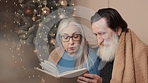 Senior couple spending happy time at home reading a book together with a decorated christmas tree