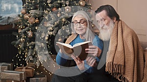 Senior couple spending happy time at home reading a book together with a decorated christmas tree