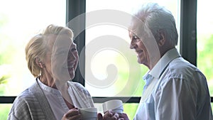 Senior couple standing near window and having coffee in living room