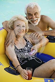 Senior Couple in Swimming Pool woman lying on inflatable raft holding drink listening to portable music player portrait.