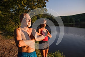 Senior couple in swimsuit standing by lake outdoors doing yoga.