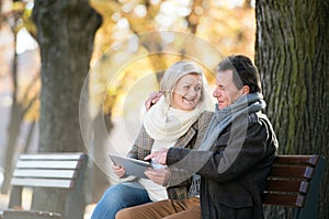 Senior couple with tablet sitting on bench. Autumn park.