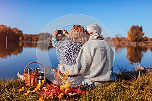 Senior couple taking selfie while having picnic by autumn lake. Happy man and woman enjoying nature and hugging