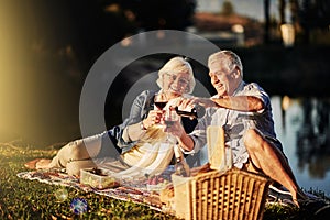 Senior couple, talking and picnic in outdoor for wine, romance and relax by lake in nature. Elderly people, celebrate