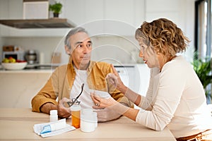 Senior couple talking and reading leaflet of drugs before drinking pills, sitting at kitchen interior