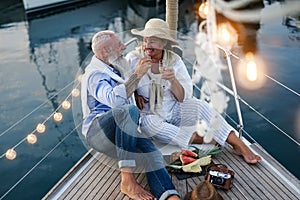 Senior couple toasting champagne and eating fruits on sailboat vacation - Happy elderly people having fun celebrating wedding