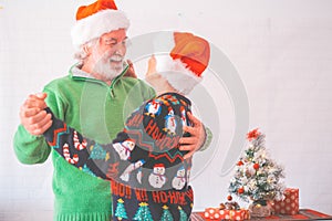 Senior couple in warm clothing and santa hat holding each others hands and dancing in front of decorated christmas tree at home.