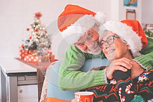 Senior couple in warm clothing and santa hat holding each others hands in front of decorated christmas tree at home. Loving old