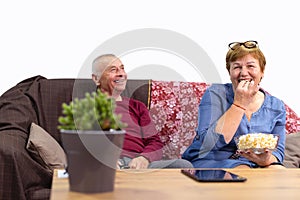 Senior couple watching tv on the sofa near table with digital tablet at home, smiling eating popcorn. Strong and