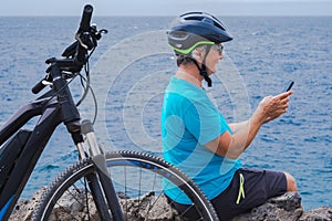 Senior cyclist woman in outdoor at sea stops on the cliff using mobile phone. Retired people with sport helmet sitting close to