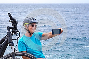 Senior cyclist woman in outdoor at sea stops on the cliff using mobile phone. Retired people with sport helmet sitting close to