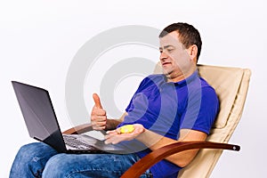 Senior deaf man holds a hearing aid in his hand and shows thumb up at the laptop camera while sitting in a chair on a