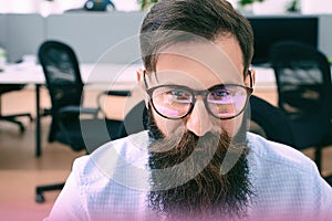Bearded man in glasses with screen reflection working on computer in IT office, sitting at desk writing code, working on