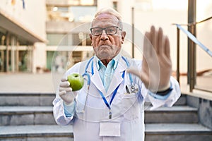 Senior doctor with grey hair holding healthy green apple with open hand doing stop sign with serious and confident expression,