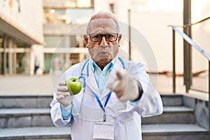 Senior doctor with grey hair holding healthy green apple pointing with finger to the camera and to you, confident gesture looking