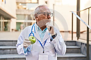Senior doctor with grey hair holding healthy green apple serious face thinking about question with hand on chin, thoughtful about