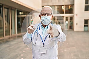 Senior doctor with grey hair wearing safety mask holding syringe pointing with finger to the camera and to you, confident gesture