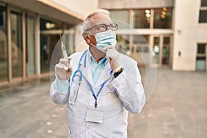 Senior doctor with grey hair wearing safety mask holding syringe serious face thinking about question with hand on chin,